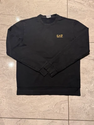 Svart EA7 sweatshirt med gul logga - Svart sweatshirt från EA7 med gul logotyp på bröstet och dekorativa gula ränder i nacken. Tröjan har rund hals, långa ärmar och ribbade muddar vid ärmslut och nederkant. Perfekt för en stilren och sportig look. Passar folk mellan S-M