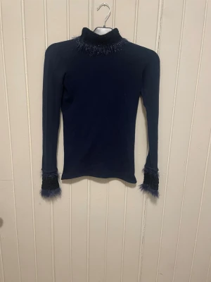 Svart polotröja med fluffiga detaljer - Snygg unik polotröja med lång ärm och unika fluffiga detaljer vid krage och ärmslut. Tröjan har en åtsittande passform och är tillverkad i ett mjukt material. Perfekt för dig som vill sticka ut med en cool och trendig look.