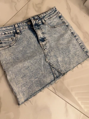 Ljusblå jeanskjol med rå kant - Trendig ljusblå jeanskjol i kort modell med rå, fransig kant nertill. Kjolen har klassisk femficksdesign, knapp och dragkedja framtill. Perfekt för dig som gillar en avslappnad och cool stil.