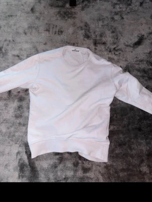 Vit tröja från Stone Island - Vit tröja från Stone Island med klassisk rund halsringning och ribbade muddar. Tröjan har den ikoniska svarta logotypen med gult broderi på ärmen. Perfekt för en clean och stilren look. SKRIV INNAN NI VILL KÖPA 