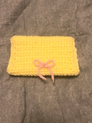 Gul virkad korthållare med rosett - Söt handgjord korthållare i ljusgul virkad design med en liten rosa rosett framtill. Den har en enkel rektangulär form och stängs med ett lock. Perfekt accessoar för dig som gillar unika detaljer och mjuka material.