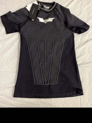 Svart Batman cosplay t-shirt - Cool svart t-shirt från YOUNGLA med Batman-motiv och rustningsinspirerat mönster i grått. Perfekt för cosplay eller maskerad. Tillverkad i stretchigt material för en tight passform. Batman-loggan syns tydligt på bröstet.