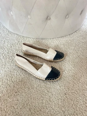 Beige och svarta ballerinaskor - Snygga ballerinaskor i beige med svart tå och flätad sula. Skorna har en klassisk design med kontrastfärgade detaljer och är tillverkade i ett glansigt material. Perfekta för en trendig och avslappnad look. Espadrillos/ Sandaler