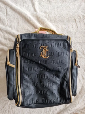 Svart necessär från Juicy Couture - Stilren svart smink väska från Juicy Couture med präglad logotyp över hela väskan och guldfärgade detaljer. Väskan har flera fack med dragkedja och ett elegant handtag i metall. Perfekt för dig som vill ha en trendig och praktisk accessoar.