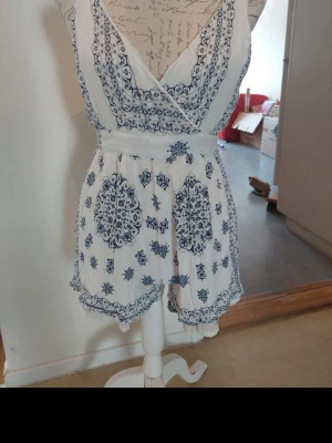 Vit och blå mönstrad jumpsuit - Supersöt vit jumpsuit med blått boho-inspirerat mönster och omlott-ringning. Kort modell med markerad midja och vida ben. Perfekt för sommardagar och festivaler. Lätt och luftig i materialet, utan ärmar och med fina detaljer.