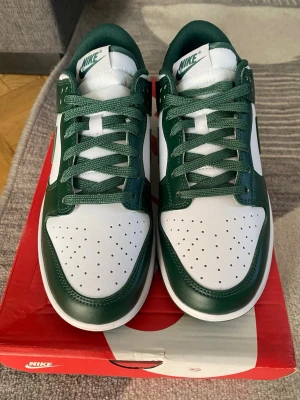 Nike Dunk Low grön/vit stl 43.HELT NYA//PRIS KAN DISKUTERAS// - Nike Dunk Low sneakers i grönt och vitt med klassisk Swoosh-logga på sidan. Ovandel i läder med perforerad tåbox och gröna skosnören. Stilren och tidlös modell som passar perfekt till streetwear och casual outfits. //PRIS KAN DISKUTERAS//