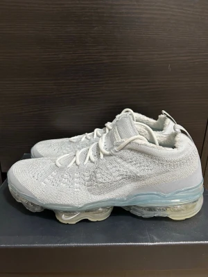 Nike Air Vapormax vita sneakers - Säljer ett par vita Nike Air Vapormax sneakers med stickad ovandel i mesh och genomskinlig sula med synliga luftbubblor. Skorna har lågt skaft, vita skosnören och diskret Nike-logga på plösen. Perfekta för dig som gillar stilrena och bekväma sneakers.