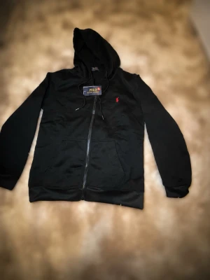 Svart hoodie från Polo Ralph Lauren - Svart hoodie med dragkedja från Polo Ralph Lauren. Klassisk design med huva och dragsko, samt röd broderad logga på bröstet. Mjuk bomullskänsla och två fickor framtill. Perfekt för en avslappnad och stilren look. Har inte använt den. Pris kan diskuteras. 