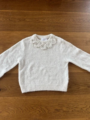 Beige stickad tröja med blommor från ZARA - Supersöt beige stickad tröja från ZARA med långa ärmar och ribbade muddar. Tröjan har dekorativa virkade blommor runt halsringningen som ger en unik och trendig look. Perfekt för dig som gillar detaljer och vill ha något extra i garderoben.