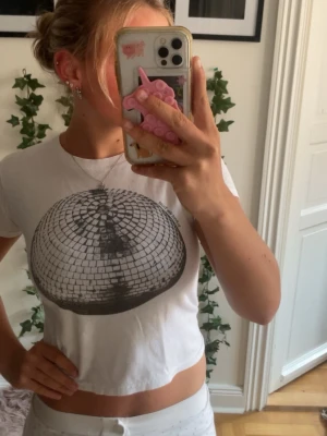 Vit t-shirt med discokula-tryck - Vit kortärmad t-shirt med ett stort tryck av en discokula på bröstet. Modellen är croppad och har en klassisk rund halsringning. Perfekt för dig som gillar en cool och trendig stil.