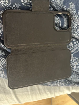 Svart Otterbox plånboksfodral till mobil, 15 Pro Max. Magsafe - Snyggt svart mobilskal i plånboksmodell med plats för kort och magnetisk stängning. Tillverkat i konstläder med en stilren design och öppning för kameran. Perfekt för dig som vill ha både skydd och funktion i ett skal. Dammig men går att rengöra på någon minut, inget slitage eller liknande.