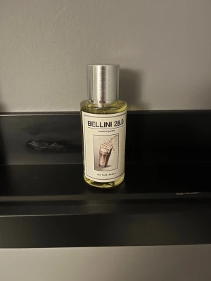 Bellini 28.0 Eau de Parfum - Bellini 28.0 Eau de Parfum från One Bold Dream i en genomskinlig glasflaska med silverfärgat lock. Etiketten har en illustration av en drink och text i svart. Doften är inspirerad av den klassiska Bellini-cocktailen.