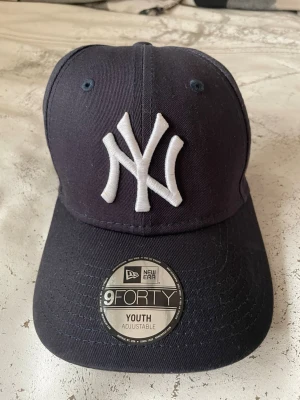 New Era Yankees 9FORTY keps marinblå - Snygg marinblå New Era 9FORTY keps med justerbar passform för ungdomar. Klassisk böjd skärm och stort vitt NY Yankees broderi framtill. Tillverkad i slitstark bomull och perfekt för dig som gillar sportig streetstyle.