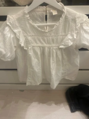 Vit blus med volanger från Gina Tricot - Söt vit blus från Gina Tricot med puffiga korta ärmar, volangdetaljer och spetsliknande band över bröstet. Blusen har rund halsringning och knäppning bak i nacken. Tillverkad i ett lätt och luftigt tyg, perfekt för en trendig look.