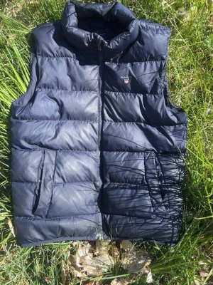 Mörkblå dunväst från GANT - Snygg mörkblå dunväst från GANT med hög krage och dragkedja framtill. Västen har två stora fickor på sidorna och diskret GANT-logga på bröstet. Perfekt att slänga över en hoodie för en chill look. Tillverkad i ett glansigt material med quiltad design.
