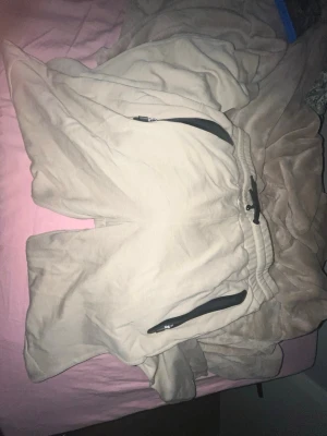 Beige mjukisbyxor med svarta detaljer - Säljer ett par beige mjukisbyxor med svarta dragkedjor på fickorna och svart dragsko i midjan. Byxorna har resår i midjan och är gjorda i ett mjukt och skönt material, perfekta för chill dagar. Passformen är relaxed och benen är raka.