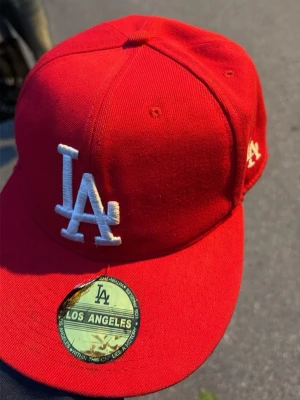 Röd LA Dodgers keps från New Era - Snygg röd keps från New Era med LA Dodgers logga broderad i vitt framtill. Klassisk flat brim och sticker kvar på skärmen. Kepsen är dock rätt sliten och kommer inte rengöra den innan försäljning för risken att stickern kan ramla av. Men den sitter ganska bra. Kepsen är inte i bra skick men helt acceptabel och inget synligt när man har på sig den. Hör av dig!