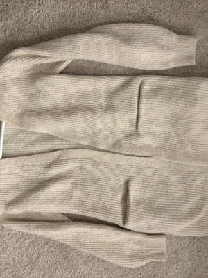 Beige ribbstickad lång kofta - Lång, öppen beige/pärlvit kofta i ribbstickat material med långa ärmar och två fickor framtill. Perfekt för lager på lager och enkel att matcha med olika outfits. Mjuk och skön känsla, passar dig som gillar en avslappnad men stilren look.