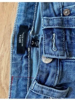 True Religion Becca Mid Rise Bootcut Jeans - Snygga blå jeans från True Religion, modell Becca Mid Rise Bootcut. Klassisk femficksdesign med dekorativa sömmar och silverfärgade knappar. Bootcut passform, perfekt under runt passar nästan till allt 