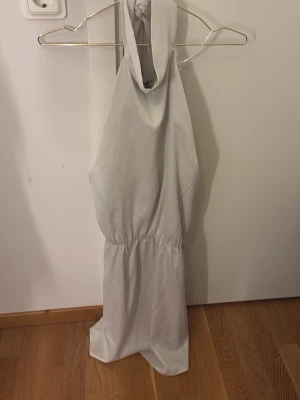 Vit halterneck jumpsuit - Snygg vit/ beige klänning med halterneck och knyt i nacken. Modellen är ärmlös och har en markerad midja för en smickrande siluett. Perfekt för dig som gillar stilrena och enkla plagg med en modern touch. Ny med prislapp.