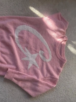 Rosa stickad tröja med stjärnmotiv - Mysig rosa stickad tröja med ett stort vitt stjärnmotiv och en virvlande linje på framsidan. Tröjan har rund halsringning och långa ärmar. Perfekt för dig som gillar gulliga och trendiga plagg med lite extra detalj.