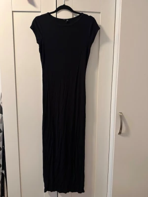 Svart ribbad långklänning - En svart långklänning med ribbad struktur och korta ärmar. Klänningen har en rund halsringning och är figurnära i modellen. Perfekt för dig som gillar en enkel men stilren look.