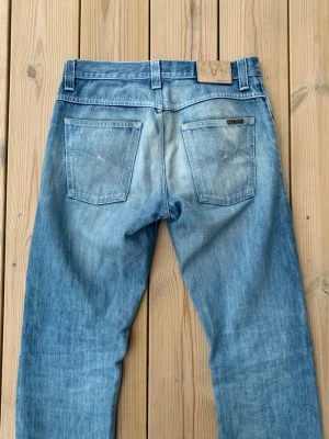 Ljusblå jeans från Nudie Jeans - Säljer ett par ljusblå jeans från Nudie Jeans med klassisk femficksmodell och raka ben. Jeansen har slitningar och en snygg tvätt som ger en avslappnad look. Tillverkade i bomull med Nudie-logga på bakfickan.