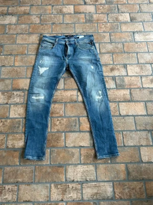 Blå Replay Broken Edge jeans - Säljer ett par blå jeans från Replay, modellen Broken Edge. Passar perfekt på dig som är ca 180 lång. De står w34 i dem men sitter som 33. Jeansen har klassisk femficksdesign, raka ben och snygga slitningar för en trendig look. Tillverkade i denim med detaljerade sömmar och patch bak i midjan. 