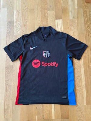 Svart FC Barcelona matchtröja Nike - Inte världens bästa kvalité på tröjan, men därav priset. Annars en fin Barcelona tröja. Bara att höra av vid funderingar. Snygg svart FC Barcelona matchtröja från Nike med klubbmärke och Spotify-logga på bröstet. Tröjan har röda och blå detaljer på sidorna och korta ärmar. Perfekt för dig som älskar fotboll och vill representera Barca på planen eller läktaren.