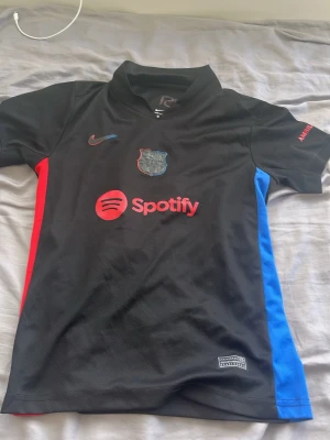 Svart FC Barcelona Nike matchtröja - Snygg svart FC Barcelona matchtröja från Nike med Dri-FIT material. Tröjan har klubbmärke, Nike-logga och Spotify som sponsor framtill samt röda och blå detaljer på sidorna. Perfekt för fotbollsträning eller att bära till vardags.