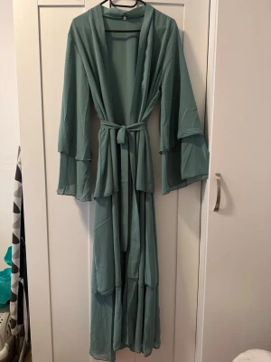 Ljusgrön långklänning med volang (abaya) - Säljer en grön långklänning med volangärmar och knytband i midjan. Klänningen har ett luftigt och flowy tyg, perfekt för varma dagar. Ärmarna är långa och har en snygg volangdetalj som ger extra stil. (Abaya)