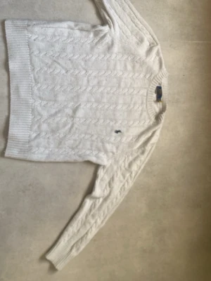 Vit kabelstickad tröja Polo Ralph Lauren - Kabelstickad vit tröja från Polo Ralph Lauren med klassisk marinblå logga broderad på bröstet. Tröjan har rund halsringning, långa ärmar och ribbade muddar. Perfekt för dig som gillar tidlös och clean stil.