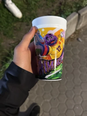 Färgglad plastmugg med Grimace-motiv - Säljer en färgglad plastmugg med motiv av Grimace i basketkläder och nummer 12. Muggen har livliga färger som lila, gul, grön och blå samt tecknad design. Perfekt för dig som gillar unika samlarsaker eller vill ha en rolig mugg till drycken.