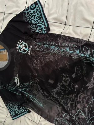 Nike Brasil svart matchtröja - Snygg svart Brasil matchtröja från Nike med turkosa detaljer och leopardmönster på ärmarna. Tröjan har ett unikt blad- och djungelmönster över hela tyget samt Brasil-logga och Nike Swoosh på bröstet. Perfekt för dig som vill sticka ut på planen.