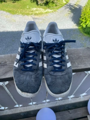 Adidas Gazelle mörkblå sneakers - Säljer ett par klassiska Adidas Gazelle sneakers i mörkblå mocka med vita detaljer och blå skosnören. Skorna har den ikoniska tre ränder-designen på sidorna och Adidas-logga på plösen. Låg modell med vit sula och guldfärgad Gazelle-text på sidan.