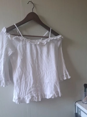 Vit offshoulder blus med volang - Söt vit offshoulder blus med volangdetaljer längs halsringningen och nederkanten. Blusen har trekvartsärm och är tillverkad i ett lätt, krinklat bomullstyg. Perfekt för en fräsch och trendig look.