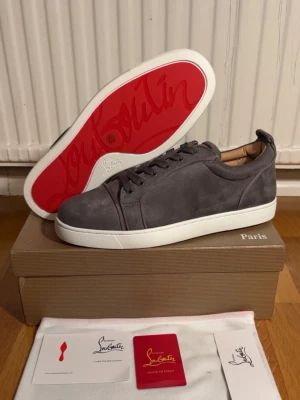 Gråa sneakers från Christian Louboutin - Stilrena gråa sneakers i mocka från Christian Louboutin med den ikoniska röda sulan. Skorna har vita gummisulor, rund tå och klassisk snörning. Insidan är fodrad i beige läder för extra komfort. Perfekta för dig som gillar exklusiva detaljer och snygg design.