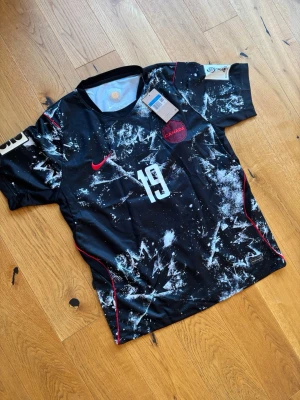 Svart Kanada fotbollströja Nike - Snygg svart fotbollströja från Nike med Kanadas landslagsemblem och nummer 19. Tröjan har vitt och rött tryck, abstrakt mönster och röda detaljer längs sidorna. På ryggen står det 'Davies' och stort nummer 19. Perfekt för dig som gillar fotboll och vill sticka ut. Helt oanvänd 