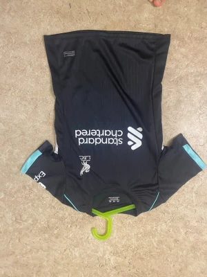 Svart Liverpool FC tränings t-shirt - Snygg svart tränings t-shirt från Liverpool FC med vita och ljusblå detaljer. Tryckt Standard Chartered-logga på bröstet och klubbmärke på framsidan. Tillverkad i ett lätt och snabbtorkande material, perfekt för träning eller sport.