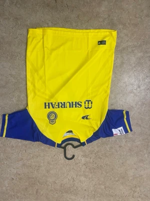 Gul och blå Al Nassr Ronaldo tröja - Snygg gul och blå fotbollströja från Al Nassr med Ronaldo och nummer 7 på ryggen. Tröjan har korta ärmar, klubbmärke och sponsorlogga framtill samt detaljer i blått på ärmar och sidor. Perfekt för dig som vill stötta ditt favoritlag på match eller träning.
