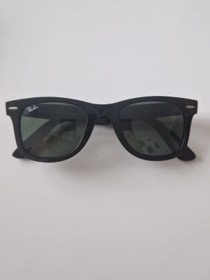 Svarta Ray-Ban Wayfarer solglasögon - Klassiska svarta Wayfarer solglasögon från Ray-Ban med ikonisk fyrkantig form och mörka glas. Snygg Ray-Ban-logga på sidan och framtill. Ramen är i plast och passar perfekt till en trendig stil.