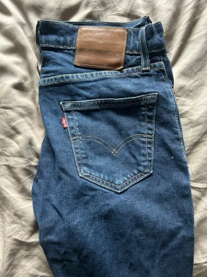 Levi's jeans - Säljer ett par klassiska blå Levi's jeans med raka ben och femficksmodell. Jeansen har kontrastsömmar, läderpatch bak i midjan och den ikoniska röda Levi's-taggen på bakfickan. Perfekta för en avslappnad och tidlös stil.