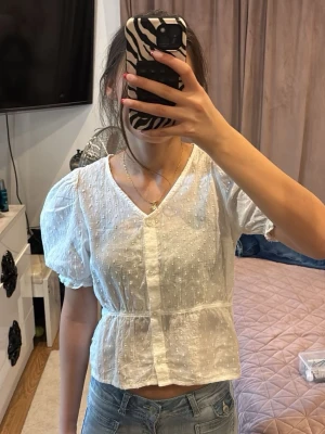 Vit blus med puffärm och v-ringning - Söt vit blus med puffärmar och v-ringning. Blusen har ett diskret prickigt mönster i tyget, markerad midja och knappar framtill. Perfekt till jeans eller kjol för en fräsch look.