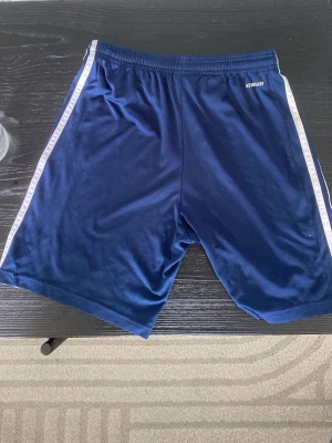 Blå Adidas shorts med vita ränder - Snygga marinblå shorts från Adidas med klassiska vita ränder längs sidorna och Adidas-logga på ena benet. Shortsen har elastisk midja och är tillverkade i ett glansigt, lätt material som passar perfekt för träning eller sport. Storlek 164/S