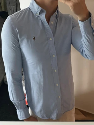 Ljusblå skjorta från Ralph Lauren - Klassisk ljusblå skjorta från Ralph Lauren med button-down krage och broderad logga på bröstet. Skjortan har lång ärm, normal passform och är tillverkad i bomull. Perfekt för en stilren och avslappnad look.