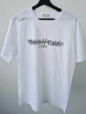 Vit Maison Margiela x Tommy Cash t-shirt - Vit t-shirt från Maison Margiela i samarbete med Tommy Cash. T-shirten har tryckt logga i svart framtill med båda märkenas namn överlappande. Klassisk rund hals och korta ärmar. Tillverkad i mjuk bomull för en clean och modern look.