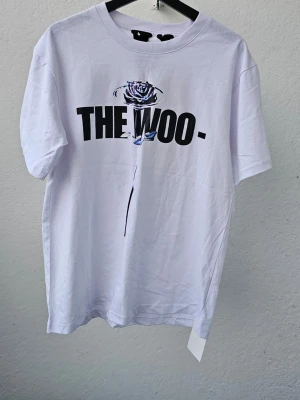Vit t-shirt THE WOO med tryck - Vit t-shirt från VLONE med svart text 'THE WOO' och en blå ros på framsidan. Baksidan har ett stort V med ros-mönster. Klassisk passform och rund hals. Perfekt för dig som gillar streetwear och snygga prints.