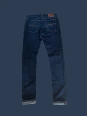 Mörkblå jeans från Nudie Jeans - Snygga mörkblå jeans från Nudie Jeans med klassisk femficksmodell och raka ben. Jeansen har kontrastsömmar och Nudie-logga på bakfickorna. Tillverkade i mjuk denim med uppvikta benslut för en avslappnad look.