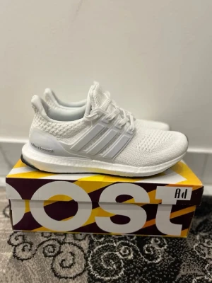 Vita Adidas Ultraboost 1.0 sneakers - Säljer ett par helvita Adidas Ultraboost sneakers med stickad ovandel och klassiska tre ränder på sidan. Skorna har en bekväm Boost-sula och snörning framtill. Perfekta för dig som gillar stilrena och sportiga sneakers..