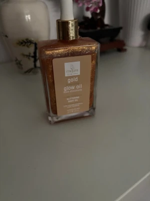 Gold Glow Oil från Majura - Gold Glow Oil från Majura är en skimrande kroppsolja i en fyrkantig glasflaska med pipett. Oljan har en intensiv guldig färg med glittereffekt som ger huden lyster. Perfekt för att framhäva glow och ge en solkysst look.